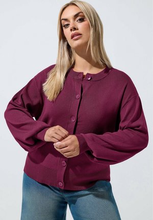 Donna con lunghi capelli biondi che indossa un cardigan prugna con bottoni e maniche lunghe e jeans blu, in piedi davanti a uno sfondo chiaro e uniforme.