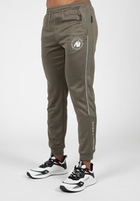Olijfgroene joggers met een elastische tailleband, witte zijpiping en een logo patch. Gepaard met zwart-witte sportschoenen.