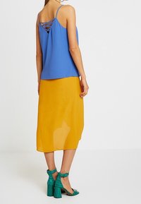 Donna con un top blu a spalline, una gonna midi giallo senape e sandali con tacco teal, in piedi di spalle contro uno sfondo neutro.