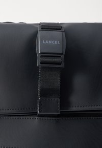 Fekete táska fényes felülettel, állítható nylon pánttal és műanyag kapoccsal, rajta "LANCEL" márkanevével. A szegélyeket varrt díszítések díszítik.