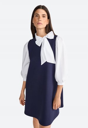 Navyblaues ärmelloses Kleid aus texturiertem Stoff mit abgerundetem Saum, kombiniert mit einer weißen Bluse mit Puffärmeln und einer großen Schleife am Kragen.