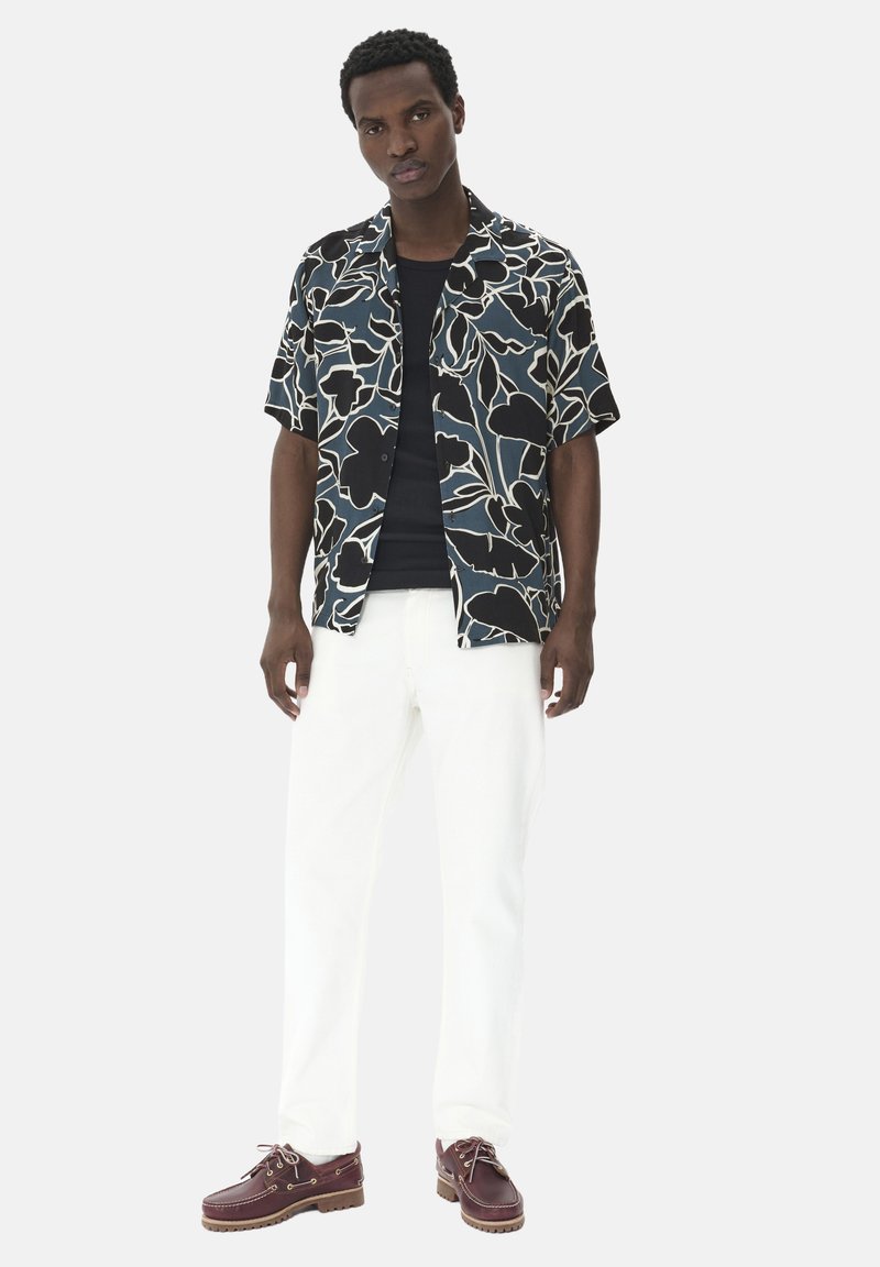 Korte mouwen bloemenpatroon shirt in zwart, wit en blauw, gedragen met witte broek en bruine bootschoenen met veters.