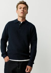Tânăr îmbrăcat cu un pulover ribbed de culoare bleumarin închis, cu guler și nasturi, și pantaloni negri, stând pe un fond deschis simplu.