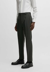 Pantaloni grigio scuro, su misura, con sottili righe verticali, tasche laterali e un design frontale piatto, abbinati a scarpe nere.