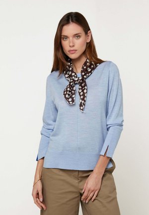 Femme aux longs cheveux bruns portant un pull bleu clair, une écharpe à pois marron et un pantalon kaki, sur un fond clair uni.