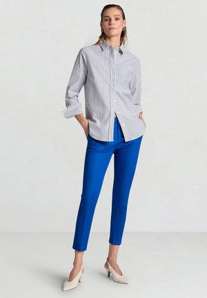 Donna in piedi con le mani nelle tasche, indossa una camicia a righe bianca e grigia con bottoni, pantaloni azzurri vivaci e tacchi beige.