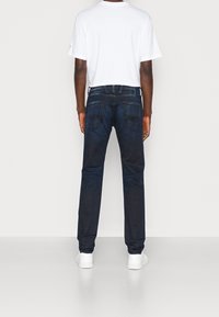 Dark blue denim jeans with a straight cut, featuring two back pockets and subtle whiskering, paired with a plain white t-shirt and white shoes.
