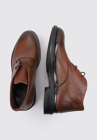 Botas de cuero marrón hasta el tobillo con punteras redondas y suelas de goma negra. Cuentan con cordones y una textura suave, diseñadas para el uso casual.