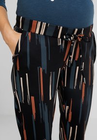 Osoba s černými kalhotami s vertikálními abstraktními pruhy v modré, rezavé a béžové barvě, se zapnutými rukama v kapsách a modrou košilí zasunutou vevnitř.