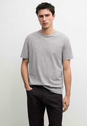 Junger Mann mit braunen Haaren, der ein schlichtes, hellgraues T-Shirt mit kurzen Ärmeln und dunkle Hosen trägt, vor einem weißen Hintergrund stehend.