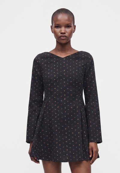 Femme aux cheveux courts portant une robe noire à manches longues avec un petit motif floral coloré et un décolleté en V.