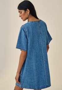Denimová šaty ve středně modré barvě, krátké rukávy, volný střih, kulatý výstřih, viditelné švy, detail s jedním knoflíkem na zadní straně.