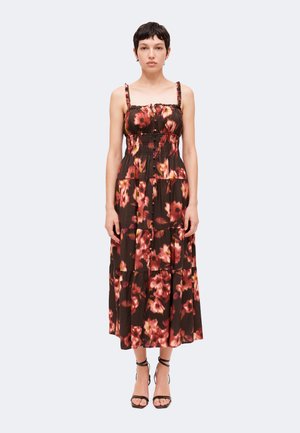 Femme debout portant une robe midi marron sans manches à imprimé floral rose et des sandales noires à talons hauts avec brides, sur un fond uni.