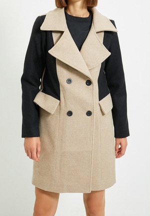 Femme portant un manteau beige et noir à double boutonnage avec de larges revers et des détails de boutons, debout devant un fond uni.