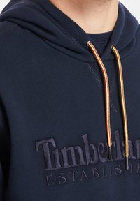 Marineblauwe hoodie met capuchon en trekkoord in het rood en geel. Voorzien van een opvallend geëtst logo in donkere stof. Zacht, gestructureerd materiaal.
