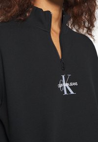 Černá mikina na zip vyrobená z měkké tkaniny, s vysokým límcem a výrazným bílým a šedým logem "Calvin Klein Jeans".