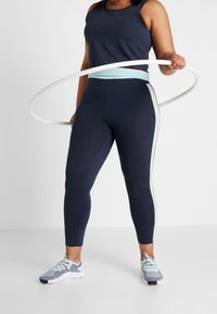Granatowe legginsy sportowe z jasnoniebieskimi paskami po bokach, zestawione z dopasowanym ciemnym topem na ramiączkach. W białym hula hoopie modelka trzyma w prawej ręce.