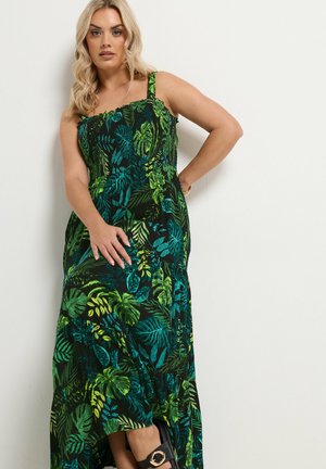 Kobieta o blond włosach, ubrana w bezrękawnikową sukienkę maxi z zielonymi i niebieskimi tropikalnymi wzorami liści, pozuje na tle białej ściany.