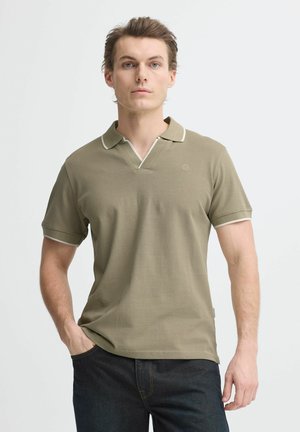 Junger Mann mit heller Haut und kurzen braunen Haaren, der ein olivgrünes kurzärmeliges Poloshirt und dunkle Jeans trägt, steht mit einer Hand in der Tasche.