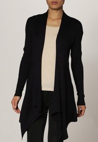 Marinblå lång cardigan i ribbad stickning med öppen framsida och draperad fåll, ihop med en ljusbeige topp och svarta byxor.