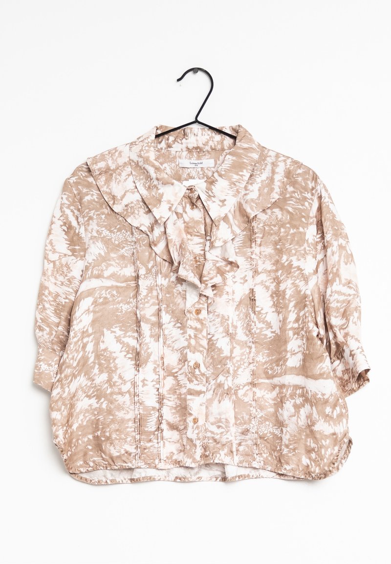 Blouse beige et blanche à motifs avec col à volants et boutons suspendue à un cintre noir sur fond blanc.