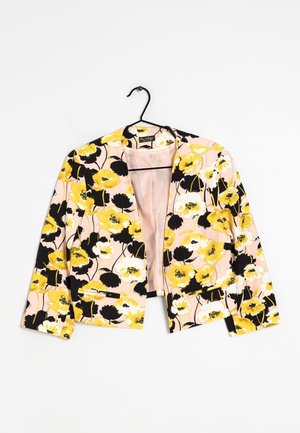 Blazer à motif floral avec des fleurs jaunes, noires et blanches sur fond rose pâle, suspendu sur un cintre noir contre une surface blanche.