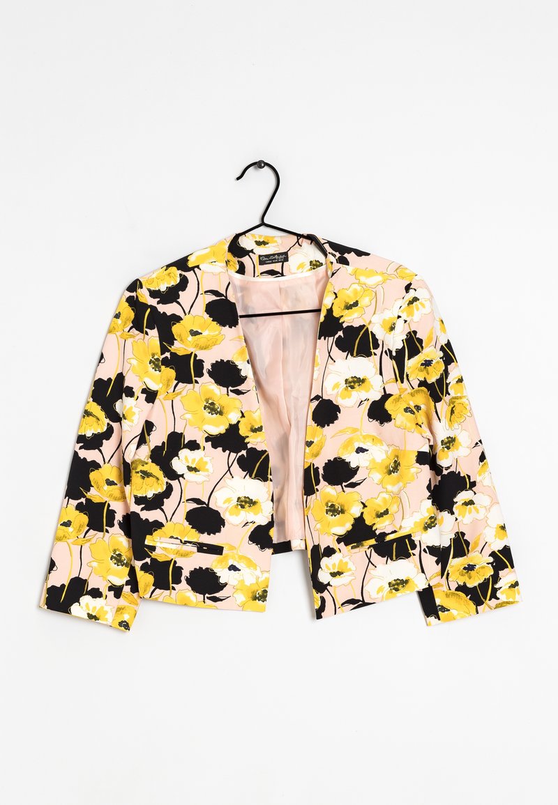 Blazer à motif floral avec des fleurs jaunes, noires et blanches sur fond rose pâle, suspendu sur un cintre noir contre une surface blanche.