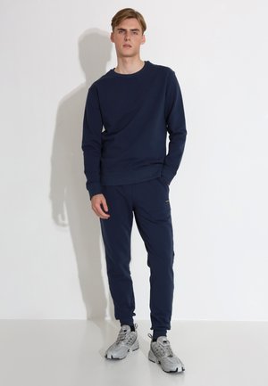 Tummansininen huppari ja sitä vastaavat verkkarit, joissa on yksinkertainen muotoilu, ribbineuloksiset hihansuut ja lahkeiden reunat, yhdistettynä harmaisiin urheilu kenkiin.