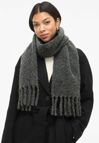 Sciarpa grigio scuro in lana testurizzata, con estremità frangiate e un design spesso e accogliente, indossata sopra un cappotto nero.