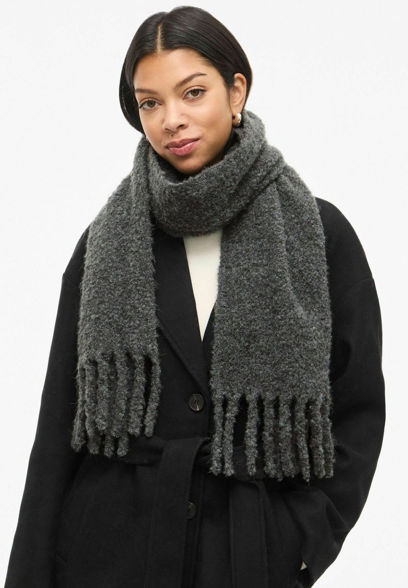 Sciarpa grigio scuro in lana testurizzata, con estremità frangiate e un design spesso e accogliente, indossata sopra un cappotto nero.