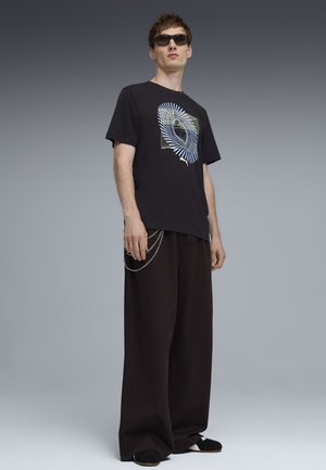 Młody mężczyzna w czarnej koszulce z nadrukiem, szerokich czarnych spodniach z dodatkiem łańcucha, czarnych wsuwanych butach i okularach przeciwsłonecznych na szarym tle.