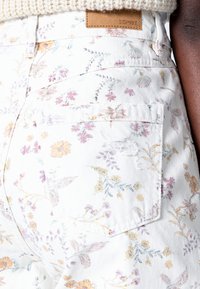 Primer plano del bolsillo de unos pantalones blancos con estampado floral, detalle desgastado y etiqueta marrón de la marca Esprit cerca de la cintura.