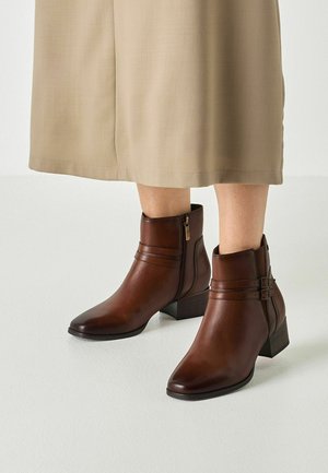 Bottines en cuir marron avec fermetures éclair et sangles latérales portées avec une jupe beige longueur mollet sur un fond de sol clair.