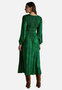 Žena v dlhých zelených kvetovaných šatách so smockovanou zadnou časťou a balónovými rukávmi, obutá v hnedých podpätkových čižmách, stojí a čelí bokom.