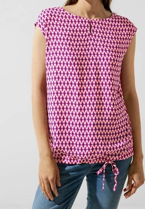Kvinde iført en ærmeløs pink og hvid mønstret top med en lille nøglehul foran og bundet kant, kombineret med blå jeans.