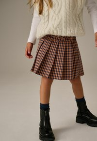 Gonna plissettata a quadri marrone e blu navy, in tessuto strutturato, abbinata a stivali neri con zip e calzini blu scuro. Gilet bianco a lavorazione a treccia indossato sopra.