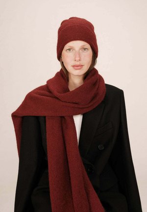 Béret tricoté bordeaux et écharpe assortie, tous deux en laine douce et texturée, portés avec un manteau noir à double boutonnage sur un haut blanc.