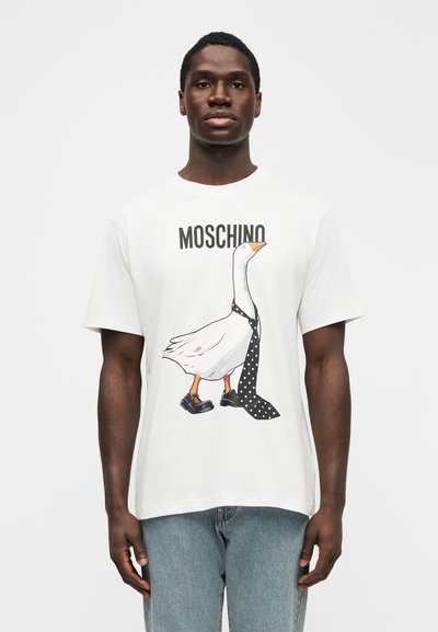 Valge T-särk, millel on kujutis hanest, kes kannab täpilist lipsu ja musti kingi. Brändinimi "Moschino" on kujunduse kohal trükitud.