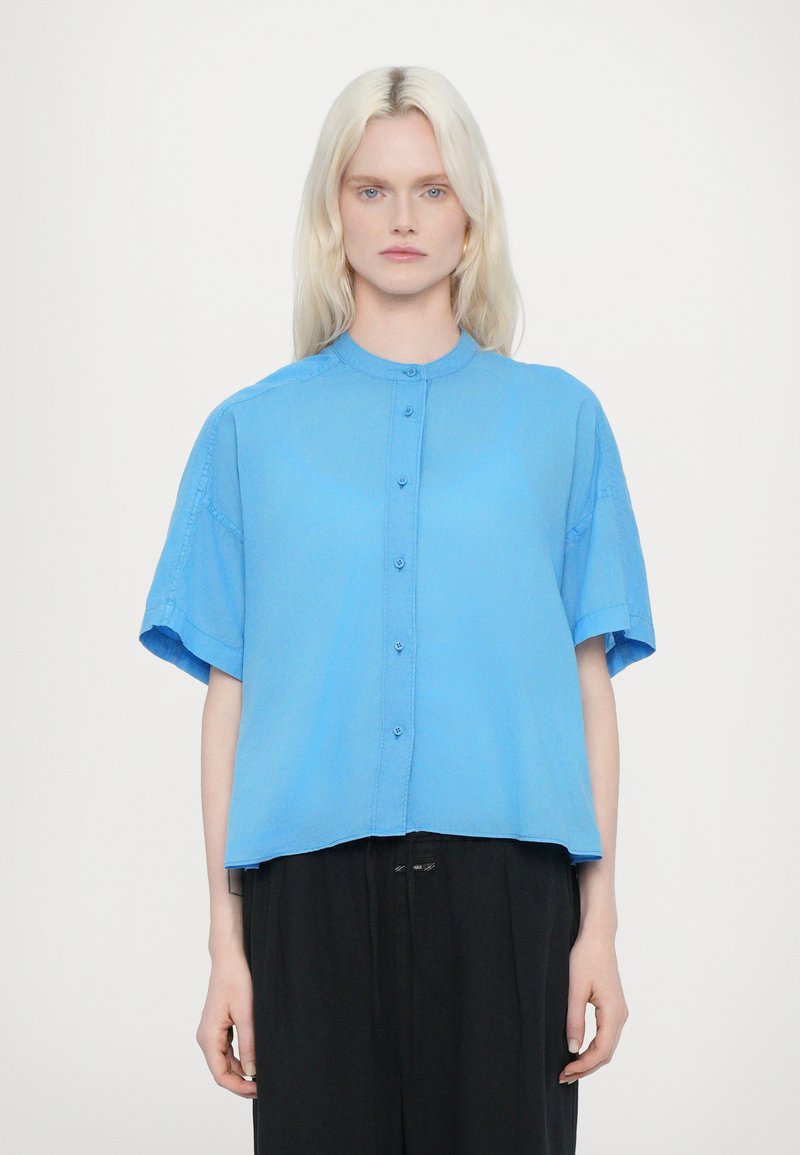 Camicia azzurra a maniche corte con colletto rotondo, caratterizzata da chiusura con bottoni frontali e vestibilità rilassata. Tessuto morbido con un design pulito.