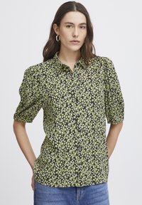 Schwarzgemusterte Hemdbluse mit Blumenmuster, die grüne und weiße Blumen zeigt. Kurze Puffärmel und weicher Stoff. 