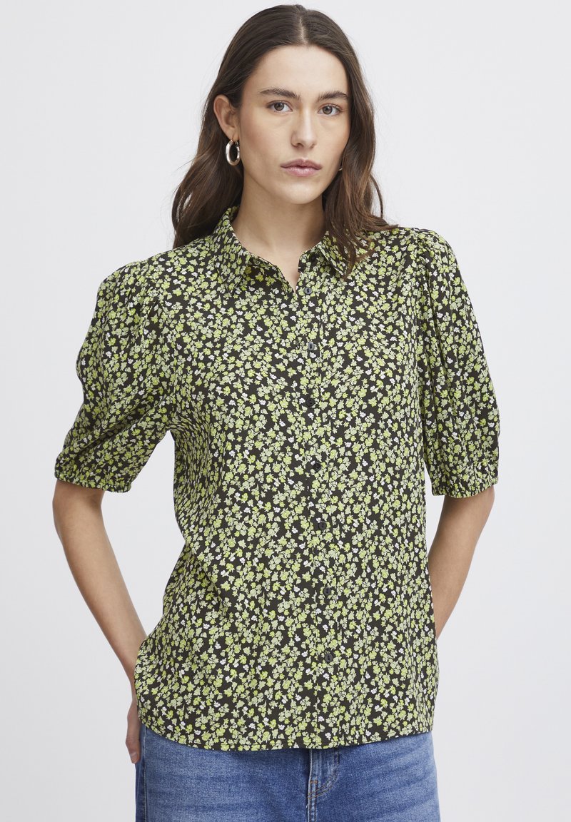 Schwarzgemusterte Hemdbluse mit Blumenmuster, die grüne und weiße Blumen zeigt. Kurze Puffärmel und weicher Stoff. 