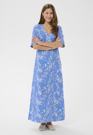 Moteris su ilgais rudais plaukais stovi sukryžiavusi rankas, dėvi mėlyną ir baltą gėlių rašto maksi suknelę ir smėlio spalvos austus butus.