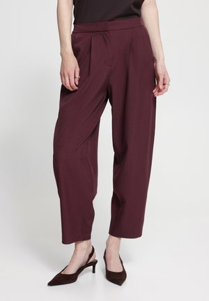 Persona che indossa pantaloni larghi color bordeaux e scarpe con tacco scure, in piedi contro uno sfondo chiaro e semplice con le mani rilassate ai lati.
