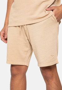 Shorts beige en mélange de coton avec taille élastique, cordon de serrage, poches latérales et logo brodé. Texture lisse et coupe décontractée.