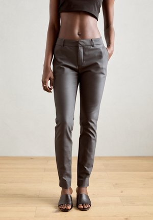 Grijs slim-fit broek gedragen door een persoon met één hand in de zak, gecombineerd met zwarte open teen slides en een cropped zwart topje.