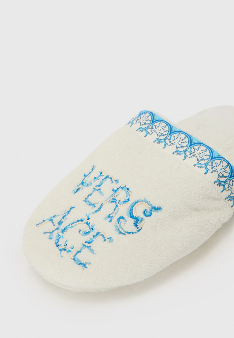 Hvide fleece slippers med blå broderet tekst "VERSACE" og dekorativt blåt mønster. Blød tekstur med afrundet tådesign.