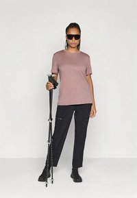 Mujer usando gafas de sol, camiseta malva, pantalones negros y botas de senderismo, sosteniendo bastones de trekking negros, de pie contra un fondo blanco liso.