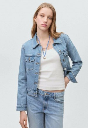Lichtblauwe denimjack met borstzakken en metalen knopen, gedragen over een witte tanktop en lichtblauwe jeans. Gladde textuur, casual pasvorm.