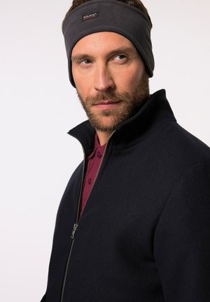 Giacca a zip in pile blu navy con colletto alto, abbinata a una fascia per capelli grigia scura; presenta una consistenza morbida e una maglietta polo bordeaux in contrasto sotto.