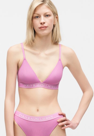 Νέα γυναίκα με ξανθά μαλλιά, φοράει ροζ bralette Polo Ralph Lauren και ταιριαστό εσώρουχο, με το χέρι στη μέση, μπροστά σε απλό φόντο.